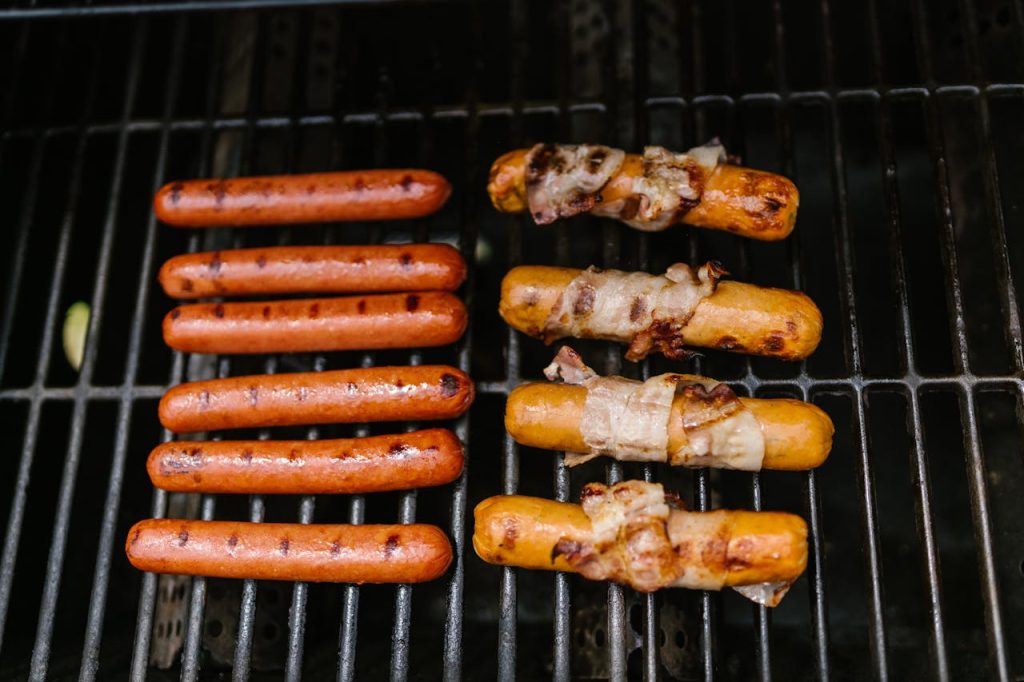 Grilled hot dogs and sausages wrapped in bacon cooking on an outdoor barbecue.